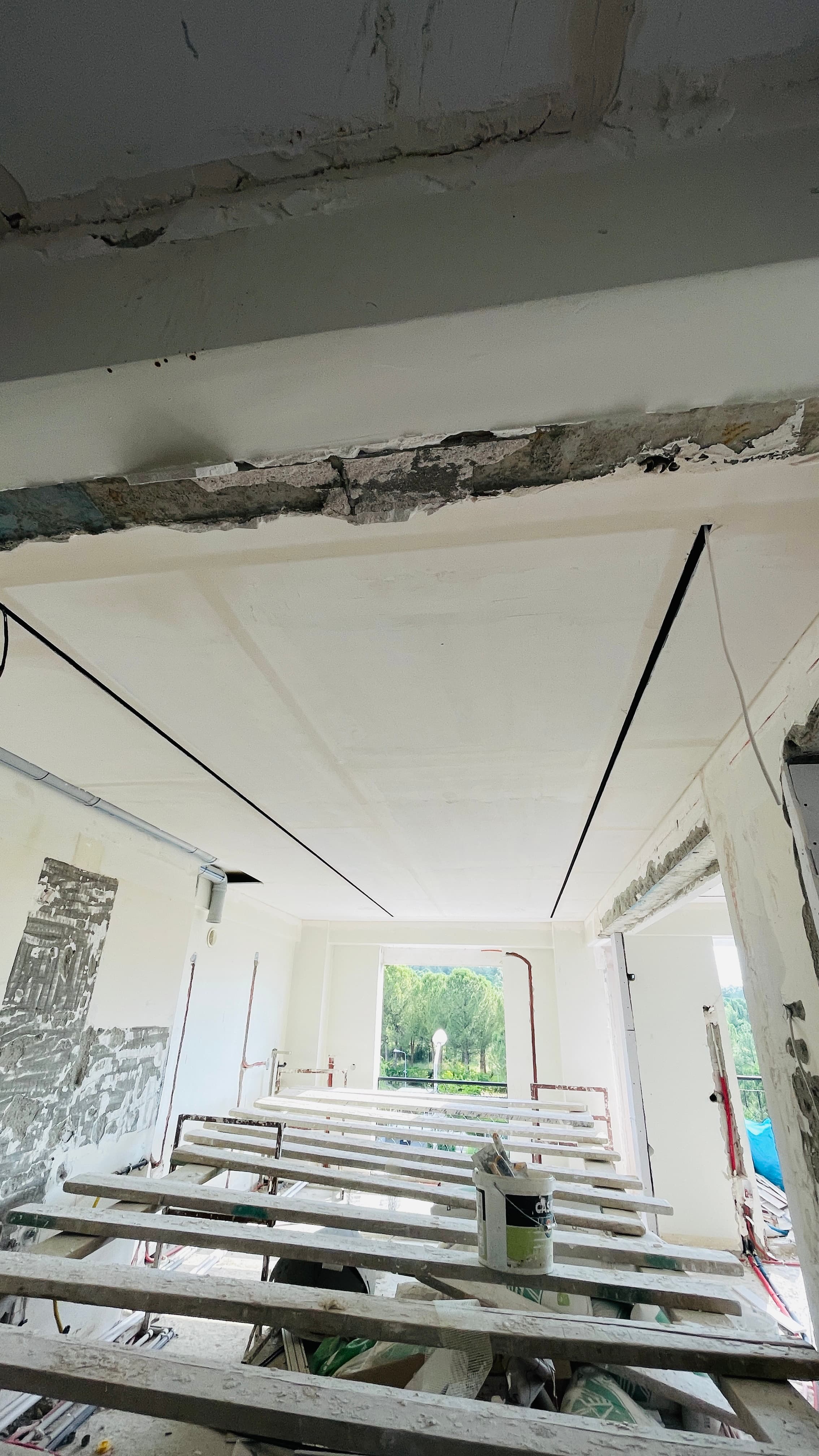 Tadilat aşamasında oda; sıva dökülmüş tavan, elektrik kabloları, ahşap iskele tahtaları, balkon manzarası.